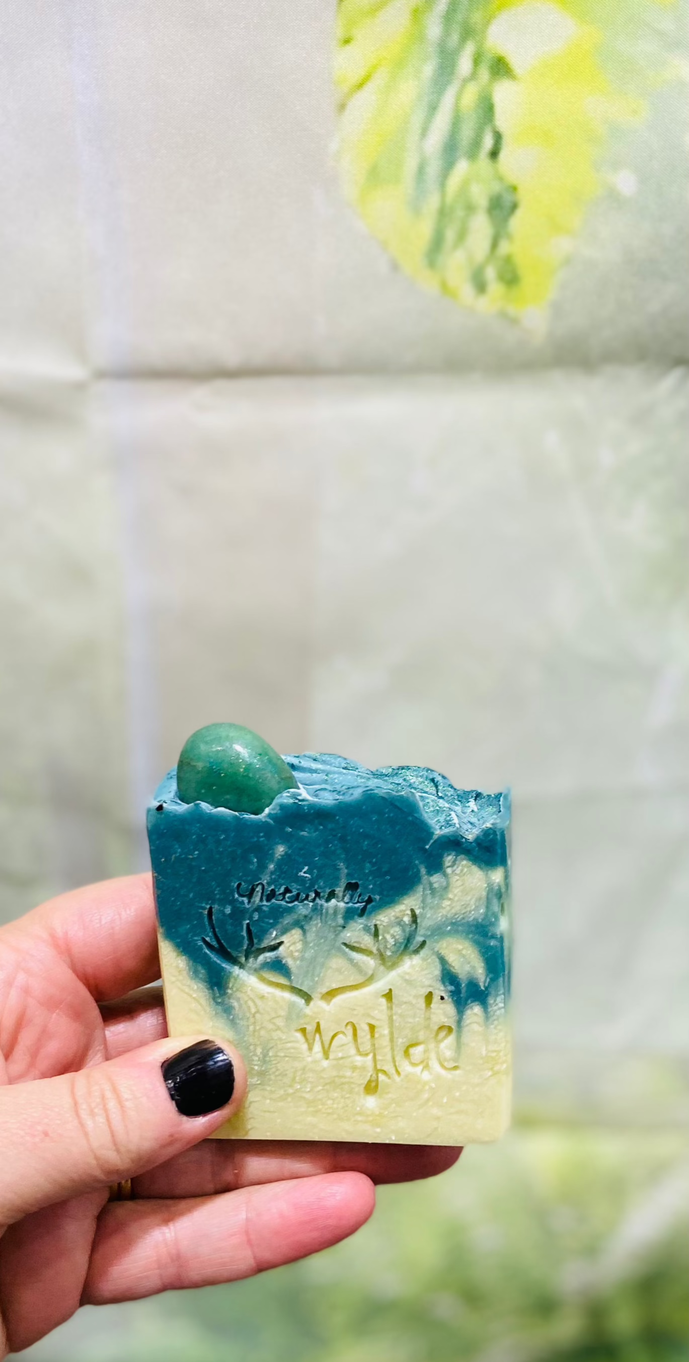Hand holding a bar of blue soap with a green stone, against a light-colored tiled wall background.