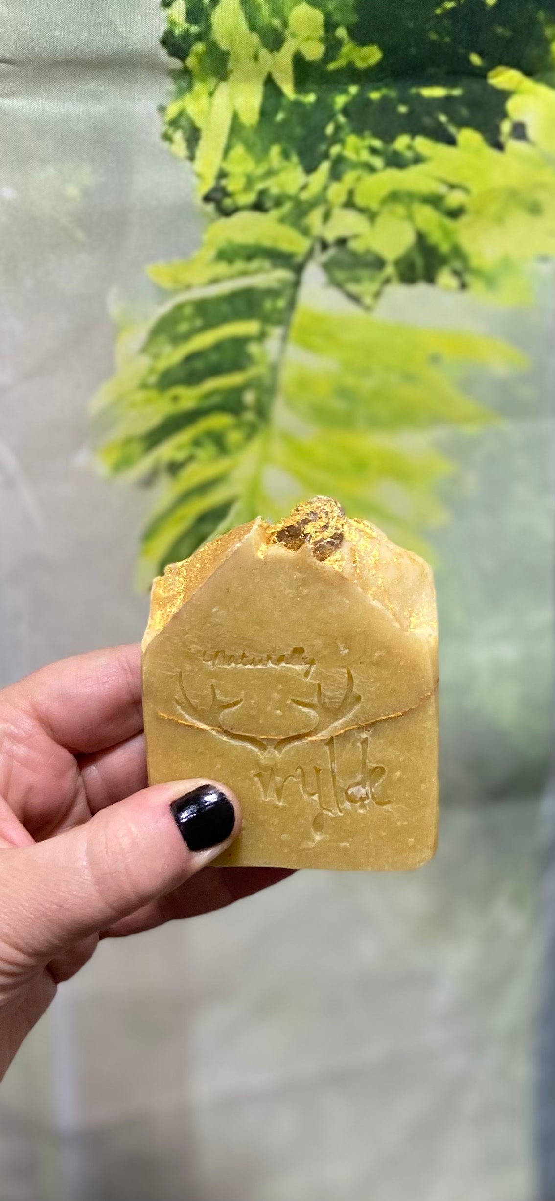 Hand holding a gold-colored soap bar with branding against a blurred green background